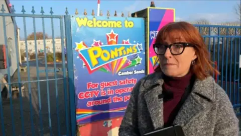 A woman with shoulder length red hair and thick black glasses stands outside the blue iron gates of the Pontins holiday park in Camber