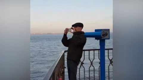 Family handout View of a man in his early thirties. He is wearing dark clothing and a cap, and has a moustache or possibly beard, but the lower half of his face is obscured by the mobile phone or camera he is holding up to take pictures with. He is standing on the corner of a seafront viewpoint, with a large telescope on a stand next to him.