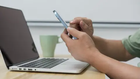 Getty Images A man using a mobile phone in front of a grey laptop placed open on a table - we can only see his arms. A green mug is in the background.