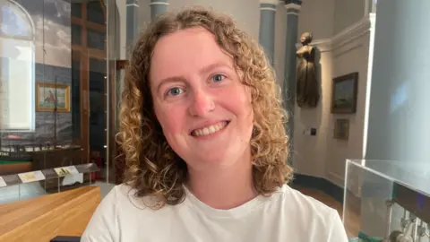 A young woman with curly hair smiling at the camera. She is wearing a white t-shirt and standing in front of a display. There area paintings on the wall behind her.