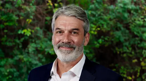 Nene Park Trust Matthew Bradbury smiles and looks directly at the camera as he is photographed outside standing in front of a green bush. He is wearing a suit jacket and white shirt, and has grey hair and a grey beard. 