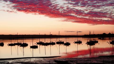 WEDNESDAY - The sunrise over Gosport with 14 small yachts silhouetted in the water in the foreground. The water in the harbour is glowing orange, reflecting the vivid red clouds above. On the horizon there are buildings and trees in silhouette against the orange sky.