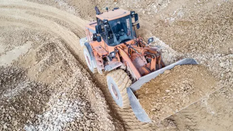 An orange digger carries a bucket full of sand and gravel. The ground it crosses in also composed of sand and gravel.