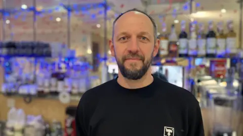 A man wearing a black jumper looks into the camera. Bottles of coffee syrup can be seen in the background.