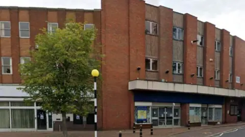 Cannock Chase District Council A three-storey building with a tree in front of it on the left of the photo, along with a black-and-white pole with orange at the top of it at a zebra crossing. 