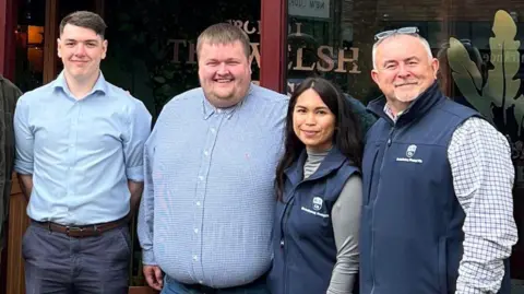 Jordan Harries Jordan stood on the left with three colleagues. They are all dressed smartly in blue attire and stood outside The Welsh House restaurant where they work.