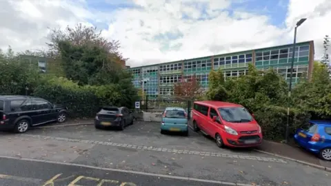 Google A green school building covered in windows stands behind a car park filled with vehicles