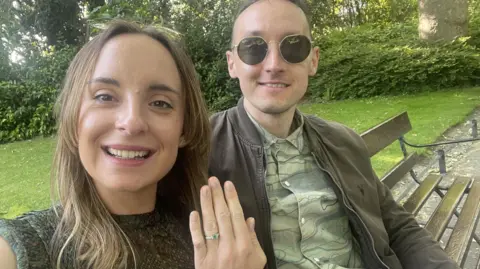 Liv Shaw (left) with long brown hair wearing a dark green sleeveless top with patterned top shows off her engagement ring with Sam (right) with short brown hair and a green shirt and brown jacket sitting next to her. They are sitting on a park bench in a park. They are both smiling.
