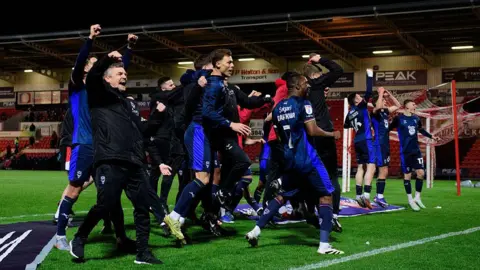 Lincoln City players and coaching staff wearing dark blue kits gather on the pitch near a goal, with several people jumping and raising clenched fists. Empty stadium seats, advertising boards and floodlit stands are in the background.