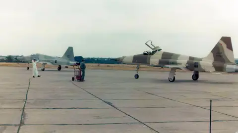 RAF Sculthorpe Heritage Centre Two F-5 Tiger II's are standing on an airbases tarmac. They are painted in an army print patten and there are two people standing near them.