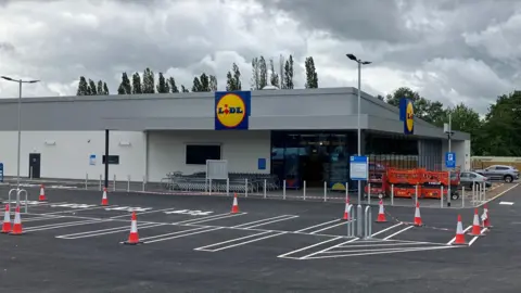 BBC A grey and white supermarket building, with an empty car park in the foreground and a large blue, red and yellow Lidl sign on the store