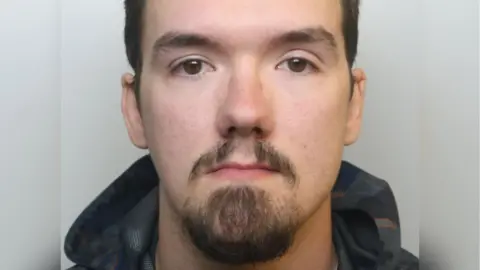 Avon and Somerset Police Jason Squibb looks at the camera in his custody picture. He has very dark eyes, dark eyebrows, dark hair and a thin moustache and goatee. He is wearing a dark raincoat. 