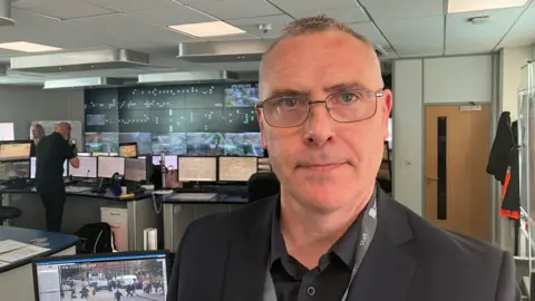 BBC / Richard Stead A man with short grey hair and glasses, wearing a black shirt and blazer, with a grey lanyard around his neck that reads 'staff', looks at the camera standing in a control room. There are monitors and computers behind him.