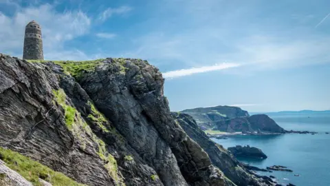 Mike Holmes Dramatic sea cliffs rising above a rugged shoreline, topped by a solitary stone tower overlooking the ocean and distant headlands beneath wispy clouds.