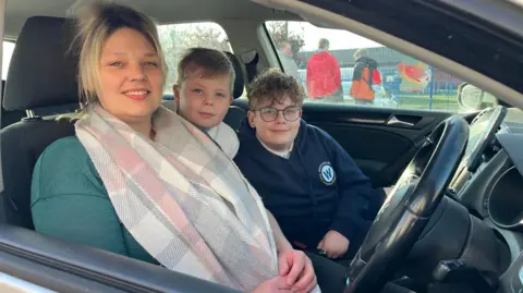 Sophie Webber is sitting in her car with her children next to her leaning into shot. She is wearing a big cream scarf and a green top. She has blonde hair. The boys are 11-year-old Freddie and 8-year-old Charlie