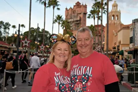 Supplied Phil Taylor has short grey hair and red t-shirt and is standing with his arm around Melanie Taylor who has long blonde hair and red t-shirt. The t-shirts are the same - with images of baubles on them along with the words 'most magical'. They are standing in an amusement park with buildings and people behind them.