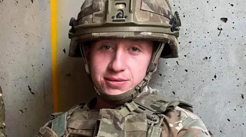 Charlie Owen is wearing camo army uniform, including a helmet, and is looking at the camera leaning against a wall