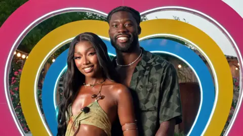 ITV /  Shutterstock Mimii Ngulube and Josh Oyinsan during the Love Island 2024 final. Mimi has long black hair and wears a strappy green dress, revealing her midriff. Josh has short braided hair and wears an unbuttoned green chequered shirt. They both smile at the camera and are pictured in front of a heart arch, made up of pink, yellow and blue hearts