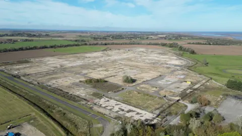 A drone view of a disused gas terminal. Little remains except roadways and hard standing. The extensive site is bordered by farmers' fields. In the distance, hedgerows and trees give way to a beach and the blue sea.