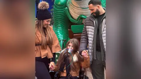 Francesca (left) with long brown hair wearing a tan jacket and black bobble hat holds the hand of her daughter, Sienna, who also has long brown hair and is wearing a tan-coloured jacket and an oxygen mask. Sienna also holds the hand of her dad, Kyle, who has black hair and beard, and a black and grey jacket and grey trousers. There is a green dinosaur in the background. They are all smiling. 