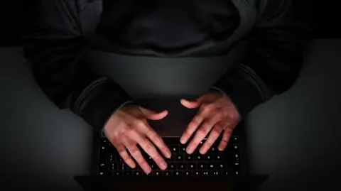 This darkened images shows a view from above of a persons hands working on the keyboard of a computer. The person is in shadow and they are wearing a black jumper. 