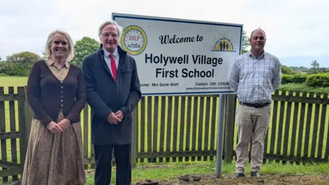School headteacher Sarah Brett with councillors Les Bowman and Guy Renner-Thompson are standing in front of the Hollywell Village First School sign. There is a wooden fence behind them with greenery in the distance.