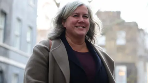 Reuters Heidi Alexander - who has long greying hair and is wearing a grey coat over a dark jacket and a purple top - smiles as she walks down the street.