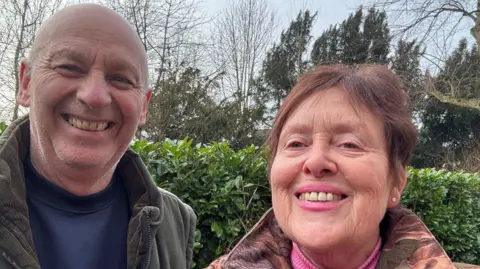 Barry and Jeana Bennion A man and a woman are smiling at the camera with a hedge behind them. Both are wearing coats. He has a blue jumper and she has a pink jumper.