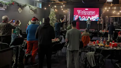 Victory Outreach Manchester Backs of guests, who are standing by tables with plates and crackers in a large room, as they watch singers and musicians perform on stage in front of a large screen saying "Welcome"