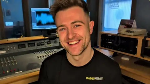Brendan Fuller smiling at the camera in a radio studio.  He has short, spikey blonde hair and is wearing a black t-shirt with a "Radio Kerry" logo on the chest.