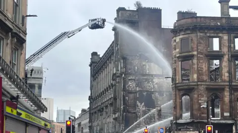 A burned out building in Glasgow, showing the damage from a fire. Fire engines spray water over the ruins.