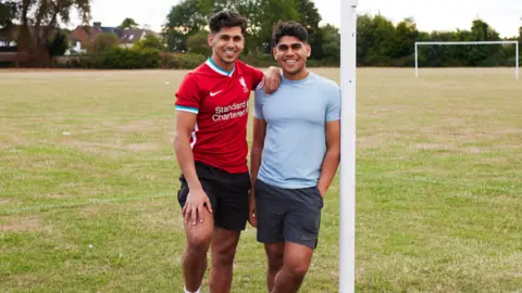 Simmer Eats Two men of South Asian heritage standing on an empty football pitch. Both of them are smiling at the camera. 