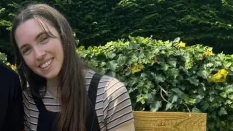 Elisha Oakley smiling for a picture outside while sitting on a bench, leaning next to her partner. He is not seen in the picture. There is some greenery behind her.