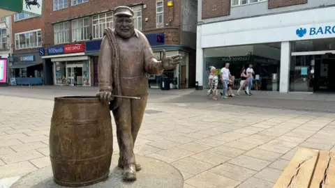 Yarn with Ernie, a bronze work in honour of local artist and potter Ernie Childs, installed in the Market Place. He is standing next to a barrel of herring.