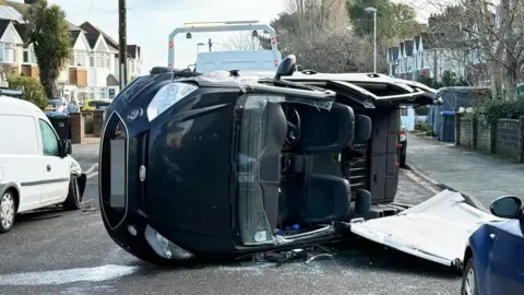 A black car is lying sideways on a road. Its roof is hanging off and its front window has almost completely come off. 
