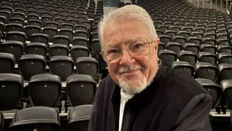 John is smiling into the camera in front of a bank of seating in the SSE Arena. He is wearing a black jacket with a white t-shirt. He is wearing large square glasses and has a hoop earring in his left ear. 