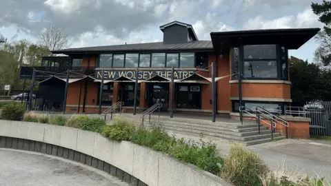 Ben Parker/BBC The New Wolsey Theatre, a red brick building with large windows and black roofs. There is planters in front with steps leading up to the building.