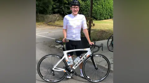 Jack stood behind his white road bike. Jack is wearing black shorts, a white cyclist top, clip-in shoes, and a helmet. He is wearing glasses and is smiling to camera.