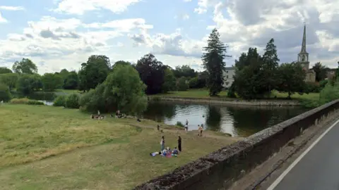 Google The River Thames at Wallingford, a grassy bank by the river where people are sat on blankets. Some are in the water. In the background is St Peter's Church, the tallest building in view, which has a pointed spire.