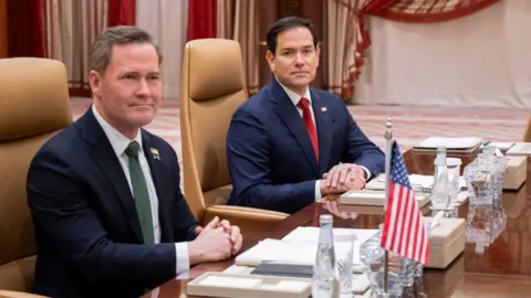 Two men in suits sit at table with their arms crossed