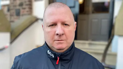 Paul Henderson stands outside Lurgan town hall. He is an older man with a shaved head. He is looking at the camera with a neutral expression on his face and is wearing a black zip up jacket.