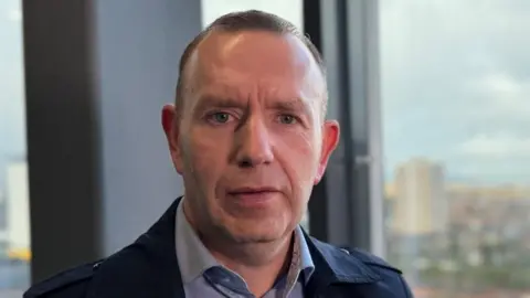 A head and shoulders image of a man wearing a blue jacket and blue shirt. He is standing in front of a window. There are buildings visible on the other side of the window.