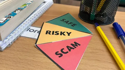 A sign that is part of the lessons is on a desk. It is clearly labelled in traffic light colours, with the words. Safe, Risky and Scam.