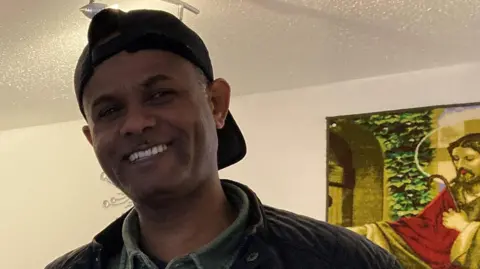 A man with a flat cap in a room stands and smiles at a camera. A portrait can be seen behind him.