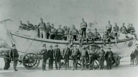 Andrew Hawkes, Poole picture archive Black and white photograph dated 1865 of lifeboat sat on a wheeled structure and crew onboard and alonside the vessel.