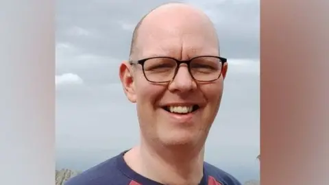 A headshot of Chris Marriott, who wears glasses and a navy t-shirt, smiling.