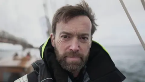 Turn to Starboard Paul Miller smiling at the camera. He has a moustache and beard and is stood on a ship.