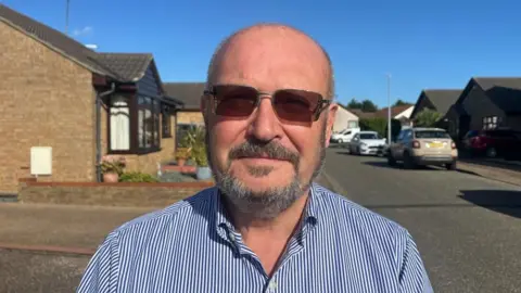 Andrew Turner/BBC County councillor Graham Plant, wearing spectacles. He has close cropped greying hair and beard with moustache. He is wearing a blue and white vertical-striped shirt and is standing in a street of bungalows and parked cars