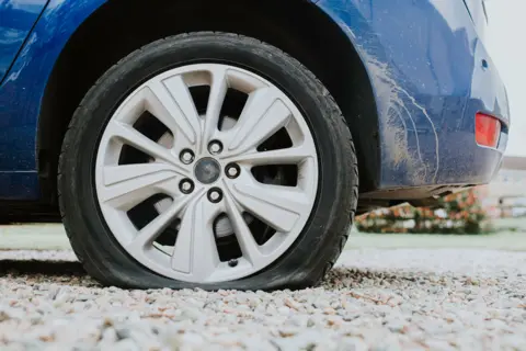 Close-up on a flat tyre on a blue car. The car is parked on a stone gravel road. The shot is taken from ground level. Most of the picture is the car, but in the background to the right hand said there is some homes, a fence and grass.