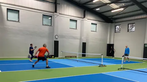 BBC Four people wearing sports gear play pickleball on an inside court which is marked in shades of blue